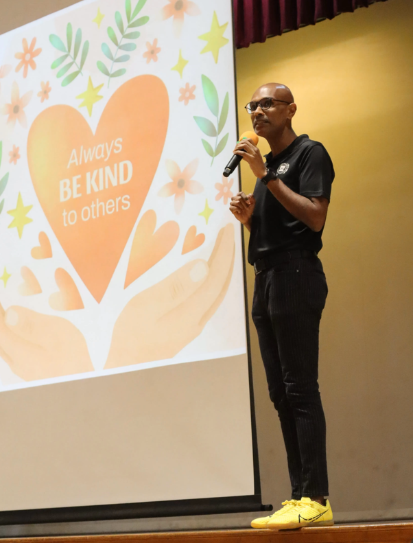 Principal speaks on stage with a projected slide reading Always BE KIND to others.